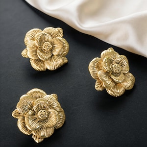 May include: Three gold-colored flower-shaped decorative objects. The objects have detailed petal textures and are arranged on a black surface with a white fabric in the background. The objects appear to be hardware or decorative accents.