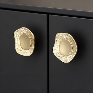 Peut inclure: Gros plan sur deux boutons de meuble dorés avec un design texturé et organique. Les boutons sont montés sur une porte d'armoire noire. Les boutons ont une zone centrale lisse de forme ovale entourée d'une bordure texturée et irrégulière.
