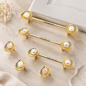 May include: A collection of gold-toned metal cabinet hardware featuring shell-shaped ends with embedded white pearls. The long handles and individual knobs are engraved with the text CHARLES. The hardware has a polished finish and a curved design.