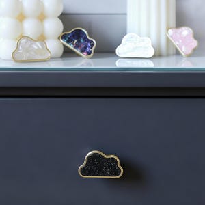K&ouml;nnte beinhalten: Wolkenf&ouml;rmige Schrankkn&ouml;pfe mit goldfarbenem Rand. Die Kn&ouml;pfe weisen verschiedene Farben und Texturen auf, darunter Wei&szlig;, schillernd und Schwarz mit Glitzer. Ein Knopf ist an einer dunkelblauen Schublade befestigt.