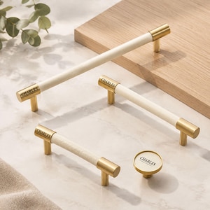 May include: A collection of cabinet hardware including three cylindrical bar pulls and one round knob. Each piece features a cream-coloured centre section with gold-toned metal ends and the word CHARLES engraved on the metal surface.