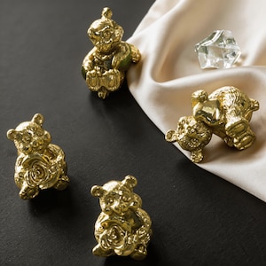 May include: Four gold-colored bear figurines. Two bears are sitting and holding a rose. One bear is sitting and holding a small object. The fourth bear is lying down. A clear, faceted crystal is also present.
