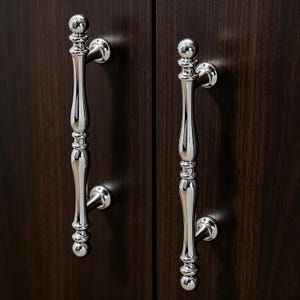 Puede incluir: Primer plano de dos tiradores de armario plateados pulidos. Los tiradores tienen un diseño decorativo y alargado con una bola en la parte superior e inferior. Están montados en una puerta de armario de madera marrón oscuro.