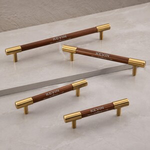 May include: Four cabinet pulls of varying lengths, each featuring a cylindrical dark brown wooden handle with gold-toned metal end caps and mounting posts. The word KEVIN is engraved in a light font on the centre of each wooden handle.
