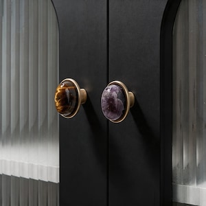 May include: Close-up of a black cabinet with two decorative knobs. One knob features a tiger's eye stone, and the other has an amethyst stone. Both knobs have a gold-colored metal base and are mounted on the cabinet doors.