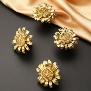 Peut inclure: Quatre boutons de meuble dorés en forme de marguerite. Les boutons ont des pétales détaillés et un centre texturé, avec une finition brillante. Le fond comprend un tissu doré et une surface sombre.