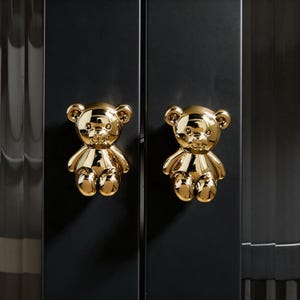 May include: Two gold-colored teddy bear cabinet knobs mounted on a black cabinet door. The knobs are highly polished and reflect light, adding a touch of whimsy to the furniture. The cabinet doors are dark and sleek.