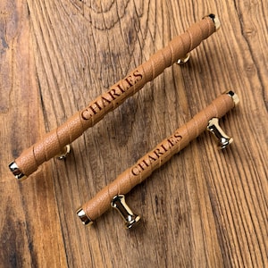 May include: Two cabinet pulls featuring a tan, textured, faux-leather wrap around a cylindrical body with polished gold-tone metal end caps and mounting posts. Each handle is engraved with the name CHARLES in a dark, sans-serif font.