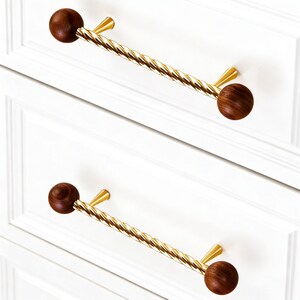 May include: Two drawer pulls with a gold twisted bar and dark brown wooden ball ends. The pulls are mounted on a white cabinet with recessed panels. The hardware has a modern, elegant design.