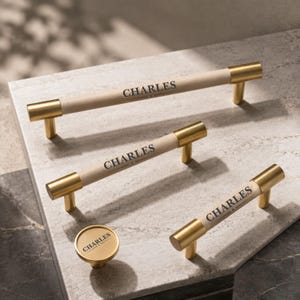 以下が含まれることがあります： マットベージュの中央部分に黒でCHARLESの文字がプリントされた3本の円筒形キャビネットプルと1つの丸型キャビネットノブ。つや消しのゴールドメタルの端部と取り付けポストが特徴の、モダンなハードウェアセットです。