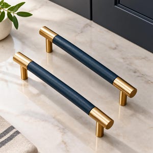 May include: Two cylindrical cabinet pulls featuring a central dark navy blue textured grip section flanked by brushed gold metal ends. Each handle is supported by two vertical gold posts, creating a linear design for furniture or cabinetry hardware.