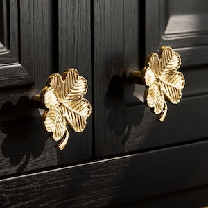 Op de afbeelding: Twee goudkleurige, bladvormige kastknoppen zijn gemonteerd op een donkere houten kast. De knoppen zijn gedetailleerd met bladnerven en stelen. De kast heeft een donkere afwerking met zichtbare houtnerf.