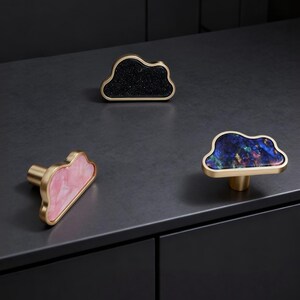 K&ouml;nnte beinhalten: Drei wolkenf&ouml;rmige Schrankkn&ouml;pfe. Einer hat eine schwarze, gesprenkelte Oberfl&auml;che, ein anderer eine rosa, marmorierte Oberfl&auml;che und der dritte eine blaue, marmorierte Oberfl&auml;che. Alle Kn&ouml;pfe haben einen goldfarbenen Metallrahmen und -schaft.