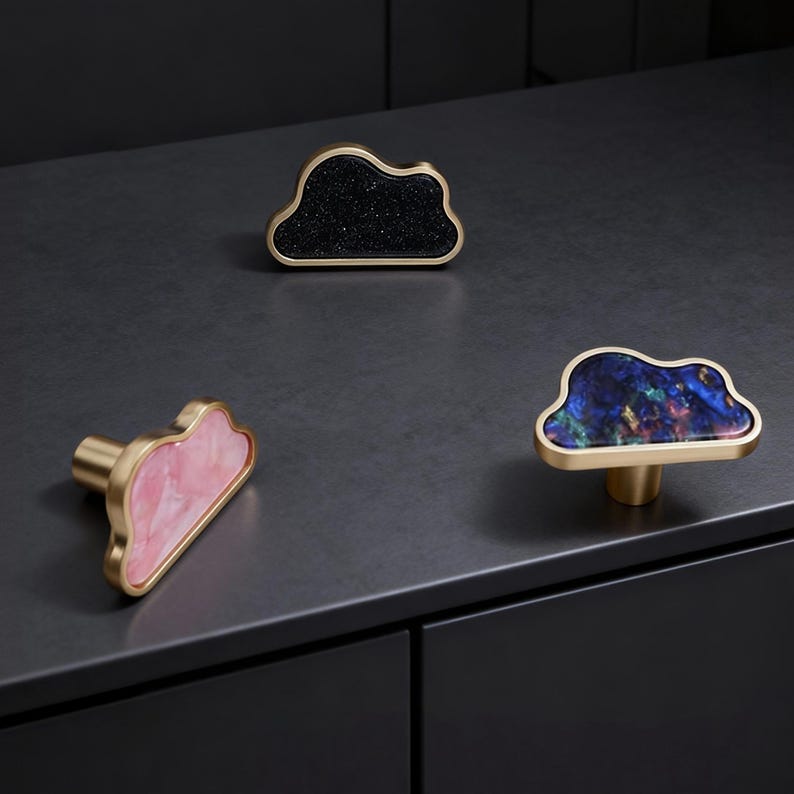 K&ouml;nnte beinhalten: Drei wolkenf&ouml;rmige Schrankkn&ouml;pfe. Einer hat eine schwarze, gesprenkelte Oberfl&auml;che, ein anderer eine rosa, marmorierte Oberfl&auml;che und der dritte eine blaue, marmorierte Oberfl&auml;che. Alle Kn&ouml;pfe haben einen goldfarbenen Metallrahmen und -schaft.