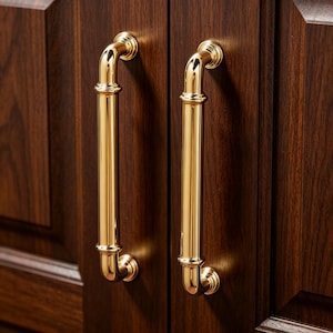 May include: Close-up of two polished gold cabinet handles against a dark brown wooden cabinet door. The handles are cylindrical with curved ends and decorative detailing, providing a classic and elegant look.