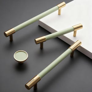 May include: Three long cylindrical cabinet pulls and one round cabinet knob, each featuring a matte sage green centre section with polished gold metal ends. The text WindmillGTDesigns is printed on the end of one long pull.