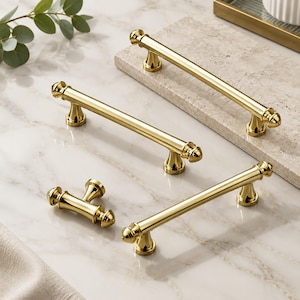 May include: A set of four polished gold-tone metal cabinet pulls and one matching T-shaped knob. Each piece features a smooth cylindrical bar design with rounded finial ends and flared bases, showcasing a reflective, high-shine metallic finish.