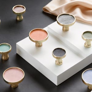 Puede incluir: Una colección de tiradores de armario de latón con tapas circulares de varios colores, incluyendo naranja, gris, verde y rosa. Los tiradores están dispuestos sobre una superficie blanca contra un fondo negro.