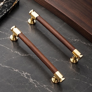 May include: Two cabinet handles featuring a cylindrical dark brown wood grip accented by polished gold-tone metal end caps and circular mounting bases. The hardware displays a smooth, straight design with metallic connectors at each end.