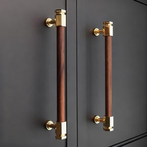 May include: Two cabinet pulls with a dark wood center and gold-colored metal accents. The pulls are cylindrical with rectangular gold end caps and circular mounting hardware. The cabinet doors are a dark gray color.