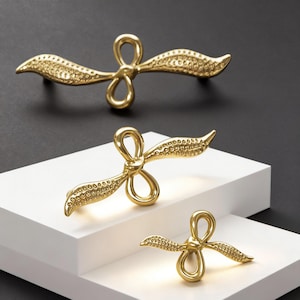 May include: Gold-coloured cabinet pulls in a bow shape. The pulls have a textured surface and are displayed on a white surface against a black background. The hardware is designed for furniture and home decor.