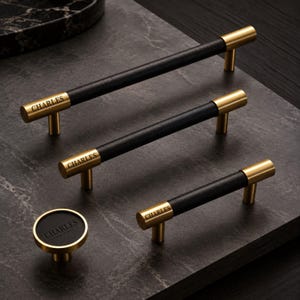 May include: Three cylindrical cabinet pulls and one circular cabinet knob featuring a black center section and brushed gold metal ends. Each piece is engraved with the word CHARLES in a serif font on the gold metal surface of the hardware.