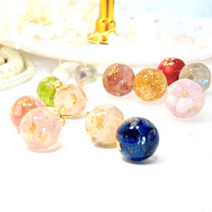 May include: A collection of colorful, round stud earrings. The earrings are made of clear resin with gold leaf and various inclusions. Colors include pink, red, green, gold, and blue. The earrings are displayed on a white surface.