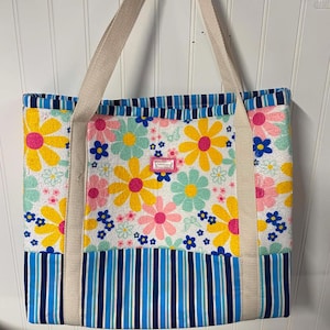 May include: A tote bag featuring a white fabric upper section with a colorful floral pattern of yellow, pink, and teal daisies, and a lower section with vertical navy blue and light blue stripes. It has two beige fabric handles and a small label.