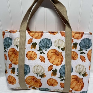 May include: A rectangular tote bag with a white background and a repeating pattern of orange, blue, and white pumpkins with brown leaves. The bag has tan handles and trim.