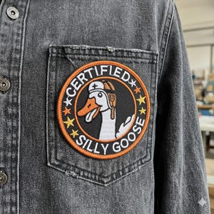 May include: A black denim jacket with a patch on the chest pocket. The patch is round with an orange border and features a cartoon goose wearing a chef's hat and the words "CERTIFIED SILLY GOOSE".