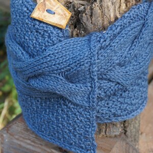 May include: A blue knitted cowl scarf with a wooden button closure. The scarf is made of a thick, textured yarn and is draped over a tree trunk. The wooden button is rectangular with a single hole.