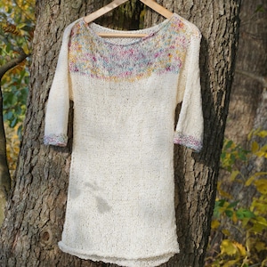 May include: Off-white knit tunic with three-quarter sleeves, hanging on a wooden hanger from a tree. The neckline and sleeve cuffs feature a colorful, speckled pattern in shades of pink, yellow, and blue. The tunic has a loose, comfortable fit.