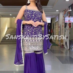 Può includere: Un completo sharara ricamato viola con una dupatta abbinata. Il top senza maniche presenta intricati ricami argentati e dorati. I pantaloni sharara hanno un design a gamba larga. Il testo "SANSKRITIBYSATISH" è visibile.