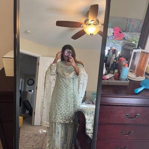 Op de afbeelding: Een lichtgroene salwar kameez set met gouden versieringen. De outfit bevat een bijpassende dupatta. Het ensemble wordt weerspiegeld in een spiegel, die de volledige outfit toont.