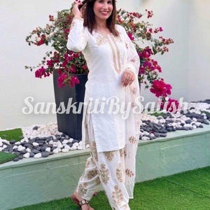 Può includere: Completo kurta e salwar ricamato bianco con stampa floreale sui pantaloni. La parte superiore a maniche lunghe ha ricami dorati intorno allo scollo e ai polsini. Il testo "Sanskriti By Satish" è visibile.