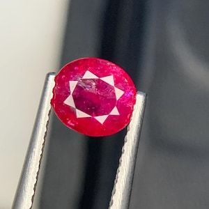 Könnte beinhalten: Ein runder, leuchtend roter Edelstein, der von einer Metallpinzette gehalten wird. Der Stein weist in der Mitte ein markantes, weißes, achtstrahliges Sternmuster auf, das sich deutlich von der tiefen, gesättigten karmesinroten Farbe abhebt.