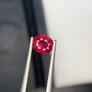 Puede incluir: Una gema roja de corte ovalado sujeta por unas pinzas de metal plateado. La piedra presenta un patrón de facetas en forma de estrella en el centro, que refleja la luz con tonos carmesí profundos y bordes claros y definidos en su superficie.