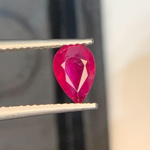 Könnte beinhalten: Ein birnenförmiger, facettierter Rubin-Edelstein mit einer leuchtenden, tiefroten Farbe. Der Edelstein wird sicher zwischen den gezackten Metallspitzen einer Pinzette gehalten und zeigt seine klaren, polierten Oberflächen und den präzisen Schliff.