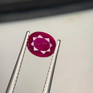 May include: An oval-shaped, deep red ruby gemstone featuring a distinct white star-shaped pattern at its center. The gemstone is held by a pair of silver-toned metal tweezers, showcasing its vibrant color and unique internal geometric design.