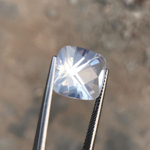 Pode incluir: Uma gema transparente, de corte quadrado, segurada por uma pinça. A gema tem múltiplas facetas que refletem a luz, criando um efeito brilhante. A pinça é prateada e o fundo é desfocado e de tom neutro.