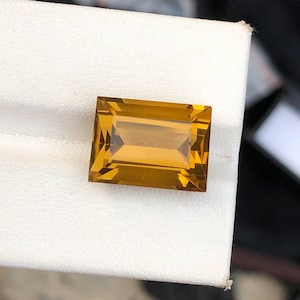 Könnte beinhalten: Ein rechteckiger, goldgelber Edelstein mit Facettenschliff. Der Edelstein ist vor einem weißen Hintergrund platziert, der seine Klarheit und Brillanz hervorhebt. Die Abmessungen des Edelsteins betragen ca. 1,9 cm x 1,3 cm.