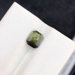 May include: A faceted, olive-green gemstone with a rectangular shape. The gem has a dark base that fades into a lighter green. The stone is set against a white, textured surface.