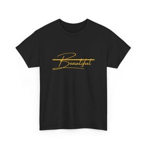 Puede incluir: Camiseta negra con la palabra "Beautiful" en dorado. La palabra está subrayada con una línea horizontal dorada. La camiseta tiene mangas cortas y cuello redondo.