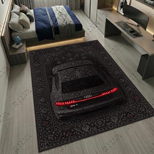 May include: A black patterned rug featuring a car design, with red taillights and the Audi logo. The rug is placed in a bedroom setting, with a bed and desk visible in the background. The rug's dimensions are not visible.