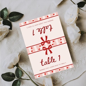 May include: A table card with the text "table 1" in red script, adorned with red bows and heart accents. The card has a light pink background with red and pink striped borders. The text "WINNERS STAY, LOSERS MOVE TO TABLE 3" is also present.