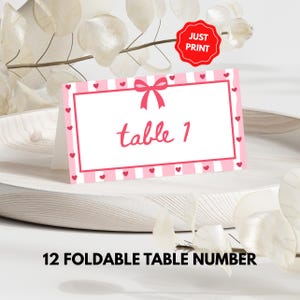 Könnte beinhalten: Eine faltbare Tischnummerkarte mit einem rosa-weiß gestreiften Rand, kleinen roten Herzen und einer rosa Schleife oben. Der Text "table 1" ist in rosa Schrift. Die Karte befindet sich auf einer weißen Oberfläche. Der Text "JUST PRINT" befindet sich auf einem roten Kreis.