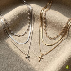 Pode incluir: Dois colares em camadas, um prateado e outro dourado, cada um com um pingente de cruz. O colar prateado tem uma corrente de cobra e uma fileira de contas transparentes. O colar dourado tem um design semelhante. Ambos os colares são exibidos em tecido bege.