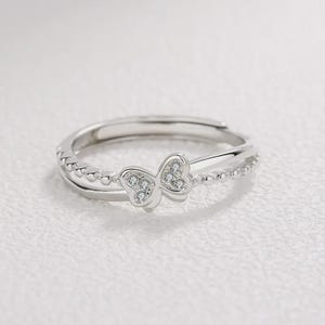 May include: A silver-colored ring with a double band design. The top band features a delicate butterfly shape adorned with small, clear gemstones. The lower band is embellished with tiny, evenly spaced beads. The ring is set against a soft, white background.