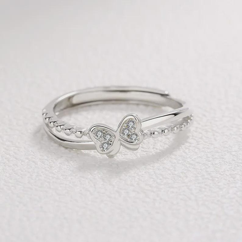 May include: A silver-colored ring with a double band design. The top band features a delicate butterfly shape adorned with small, clear gemstones. The lower band is embellished with tiny, evenly spaced beads. The ring is set against a soft, white background.