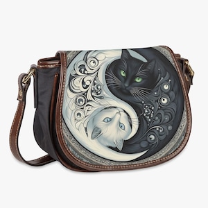 Puede incluir: Bolso bandolera negro y marrón con un diseño de Yin y Yang con dos gatos. Uno es negro con ojos verdes, el otro blanco con ojos azules. El bolso tiene una correa y ribetes de cuero marrón.